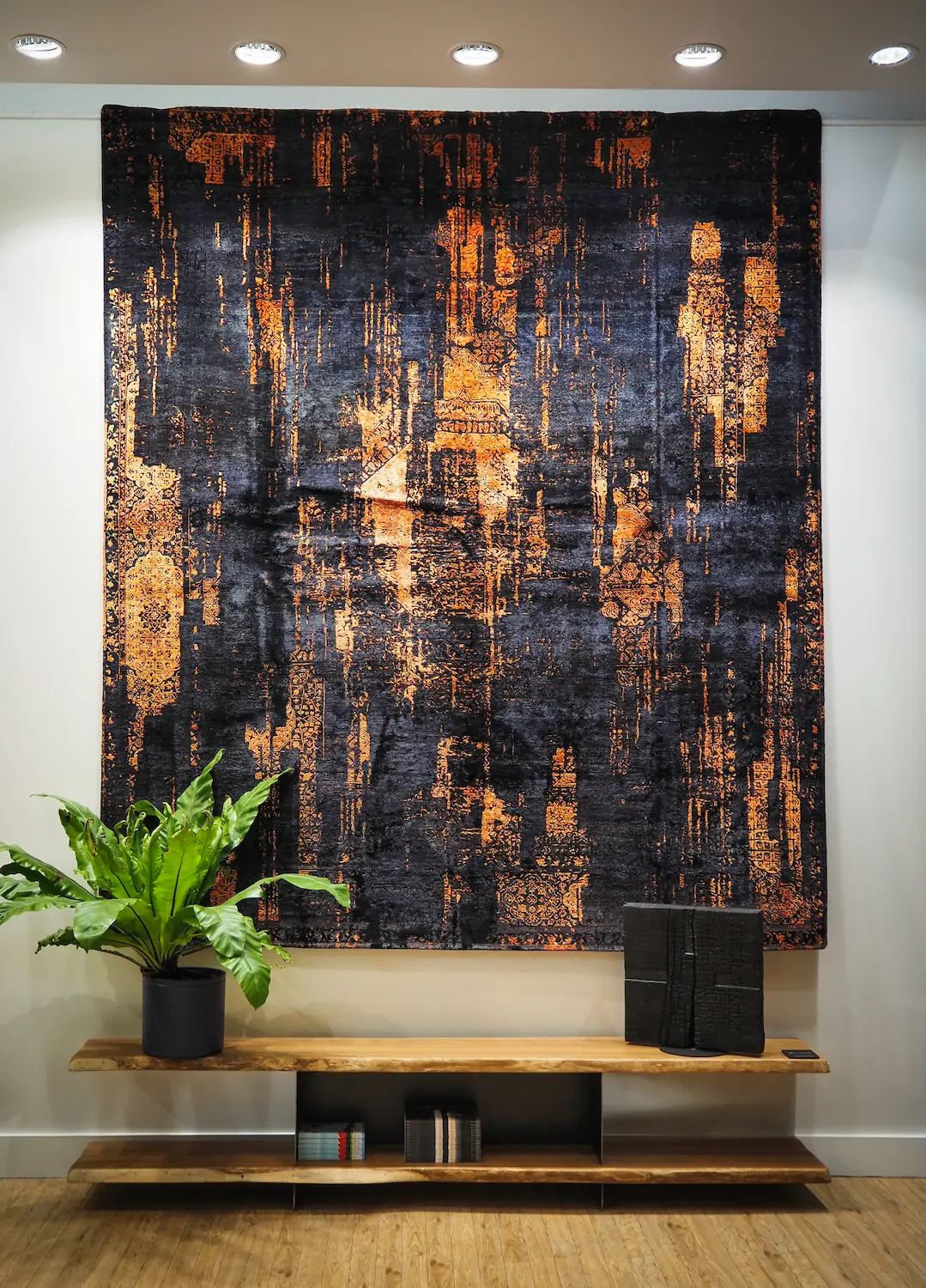 Tapis moderne noir et or – décoration murale contemporaine Grand tapis moderne noir et orange accroché au mur dans un espace de présentation avec étagère en bois et plante verte