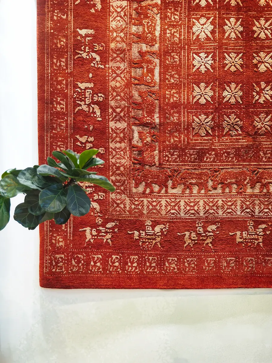 Détail tapis Pazyryk rouge – bordure traditionnelle et motifs anciens Détail coin tapis Pazyryk rouge bordeaux avec bordure animaux et motifs floraux, plante verte au sol